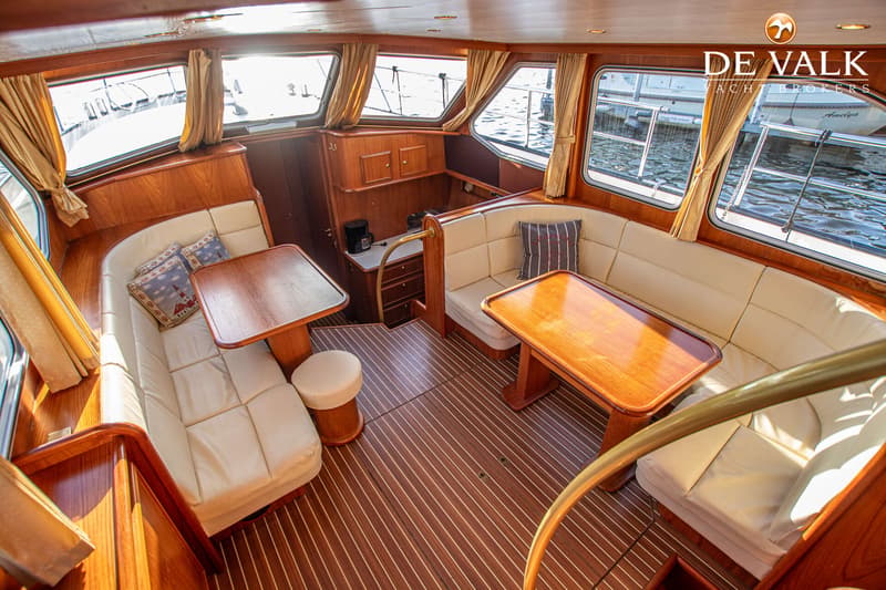 De foto toont het interieur van een boot met houten lambrisering, twee zithoeken met crèmekleurige kussens en tafels, en…