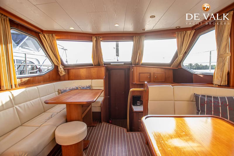 De foto toont het interieur van een boot met houten lambrisering, crèmekleurige banken, tafels en ramen met gordijnen.