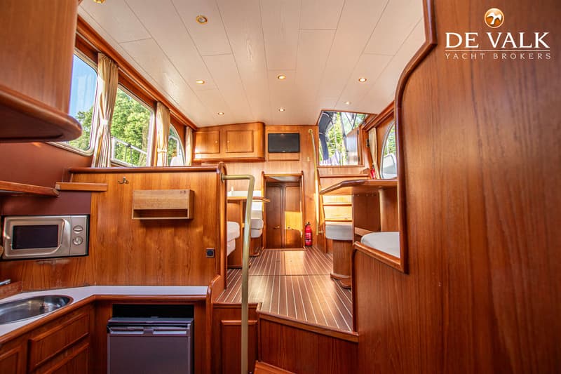 Een interieur van een boot met houten wanden, een plafond met inbouwspots, ramen met gordijnen en een keukenblok.