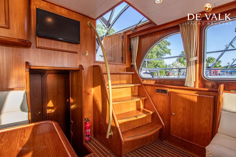 Een interieur van een boot met houten wanden, een trap, een televisie en ramen met uitzicht op bomen en andere boten.