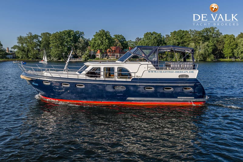 Een blauw-witte motorboot met rode onderkant vaart op het water, met bomen en huizen op de achtergrond. — De Drait Deluxe 42