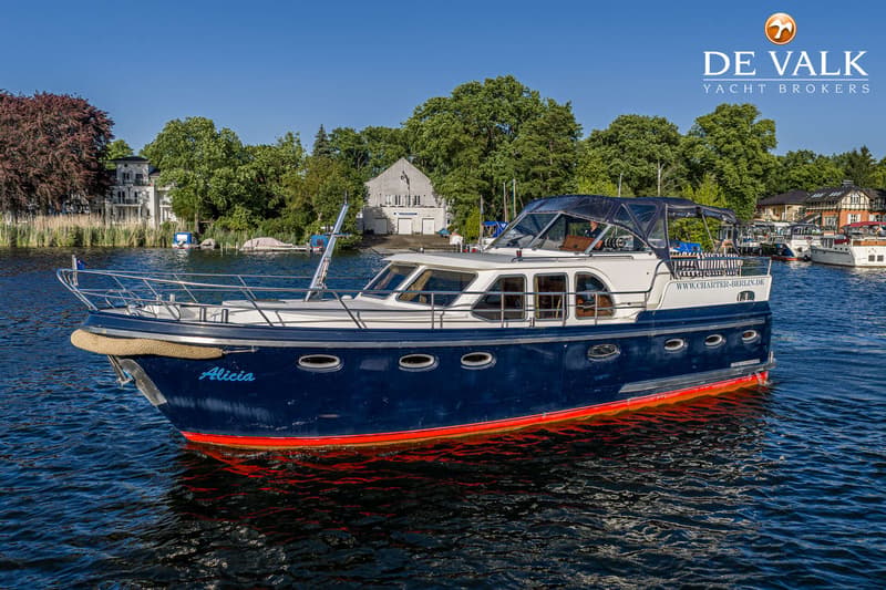 Een blauw-witte motorboot met rode onderkant en de naam "Alicia" vaart op water met een groene oever op de achtergrond.