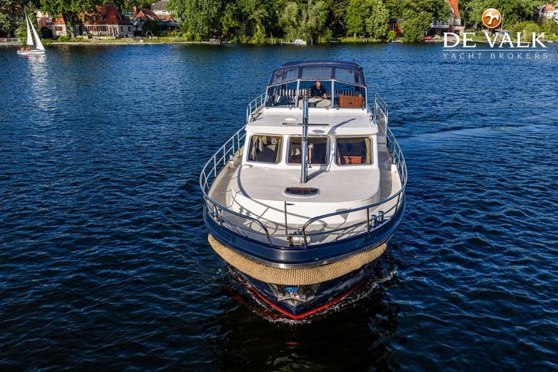 Een blauw-witte motorboot met een kapitein aan het roer vaart op rustig water, met een zeilboot en huizen op de achtergrond.