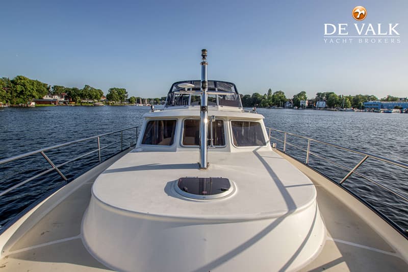 Een witte motorboot vaart op rustig water, met een landschap van bomen en huizen op de achtergrond onder een heldere blauwe…