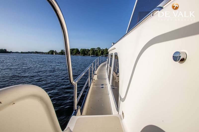 Een witte boot met een reling en ramen vaart op een meer met bomen aan de overkant onder een blauwe lucht.