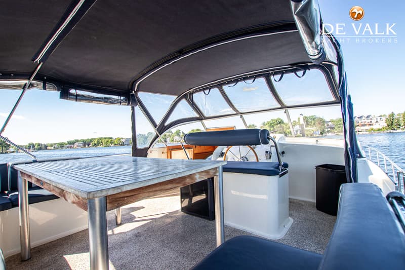 Een bootinterieur met een houten tafel, stuurwiel, blauwe kussens en een zwarte biminitop, met uitzicht op water en…