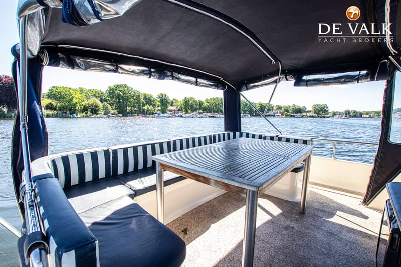 Een houten tafel met metalen poten staat op een bootdek met gestreepte en effen donkerblauwe kussens, met water en bomen op…