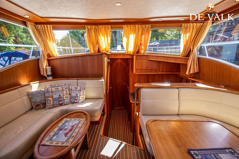 De foto toont het interieur van een boot met houten lambrisering, beige banken, een houten vloer en ramen met oranje…