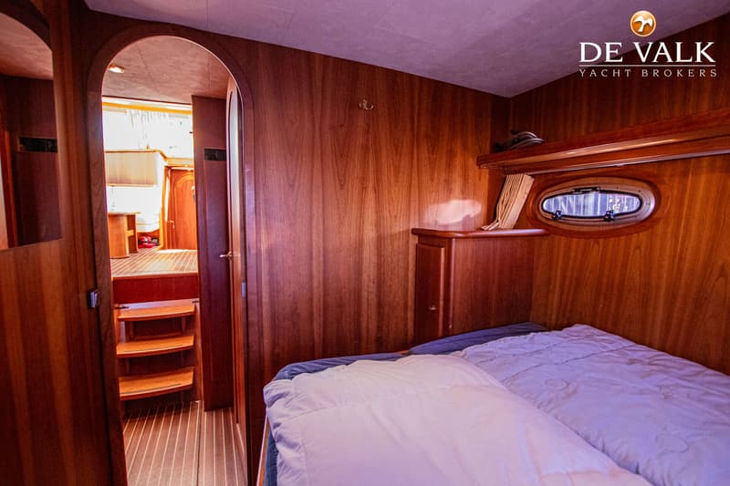 Een slaapkamer in een boot met een bed, een patrijspoort, houten wanden en een doorgang naar een andere ruimte.