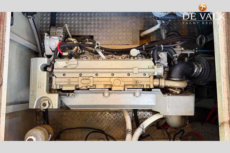 Een close-up van een scheepsmotor met diverse slangen en bedrading, geplaatst op een metalen vloer met ruitpatroon.
