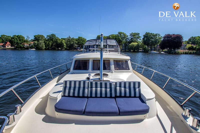 Een witte boot met blauw-wit gestreepte en effen blauwe kussens op het voordek, varend op een waterweg. — Babro Newline 1290