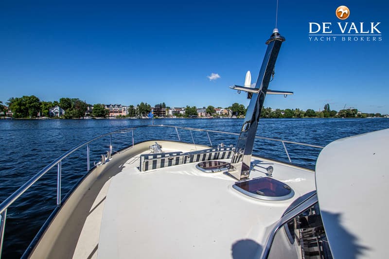 De foto toont het voordek van een boot met reling, twee patrijspoorten en een mast, varend op water met bebouwing op de…
