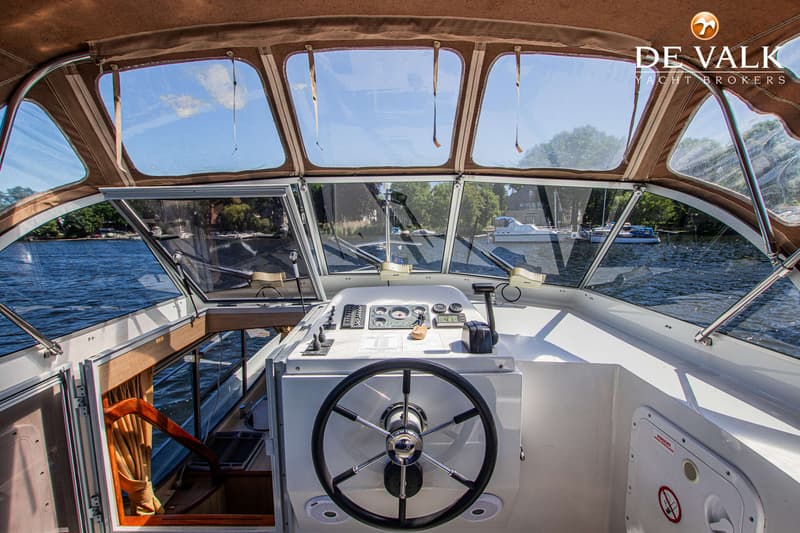 De foto toont de stuurstand van een boot met een stuurwiel, instrumentenpaneel en bedieningshendels, met uitzicht op water…