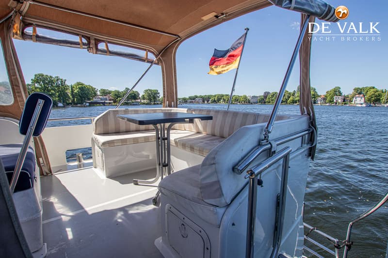 Een bootdek met zitbanken, een tafel, een Duitse vlag en uitzicht op water en beboste oever.