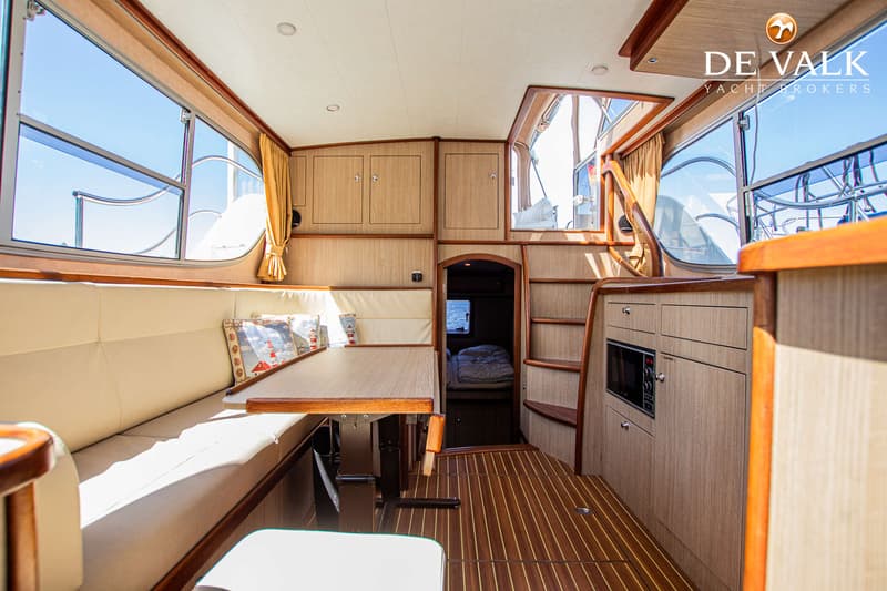 De foto toont het interieur van een boot met een zithoek, tafel, ramen, kasten en een doorgang naar een slaapcabine.