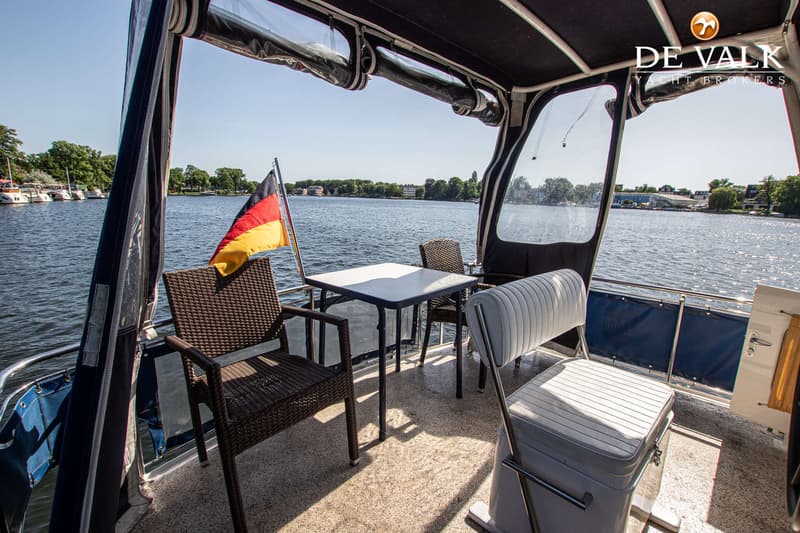 Een bootdek met een tafel, twee stoelen, een Duitse vlag en een stuurstoel, met water en bomen op de achtergrond.