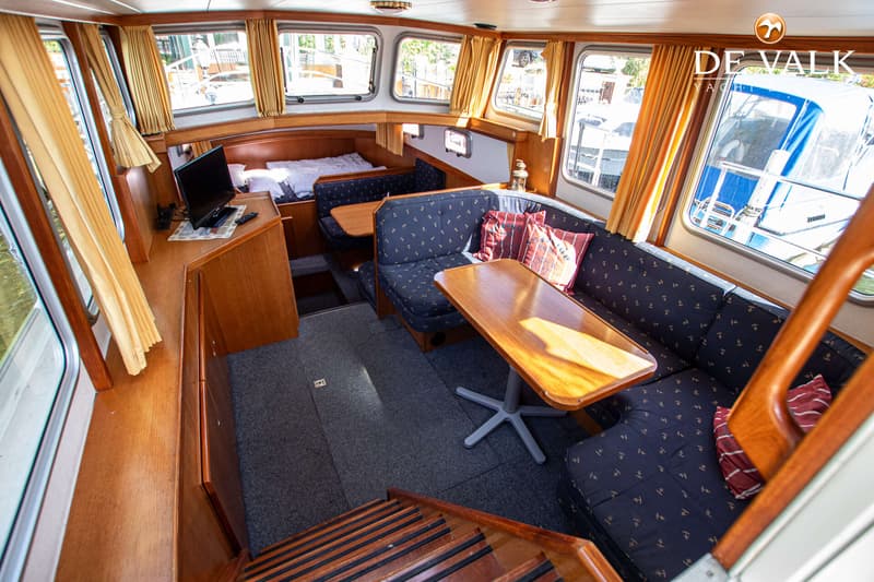De foto toont het interieur van een boot met houten afwerking, blauwe banken, een bed, een televisie en ramen met gordijnen.