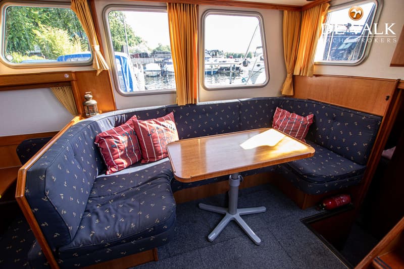 Een interieur van een boot met een U-vormige bank, een houten tafel en ramen met uitzicht op water en andere boten.