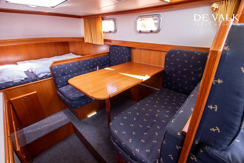 De foto toont een interieur van een boot met een houten tafel, twee banken met ankerpatroon bekleding, een bed en twee…