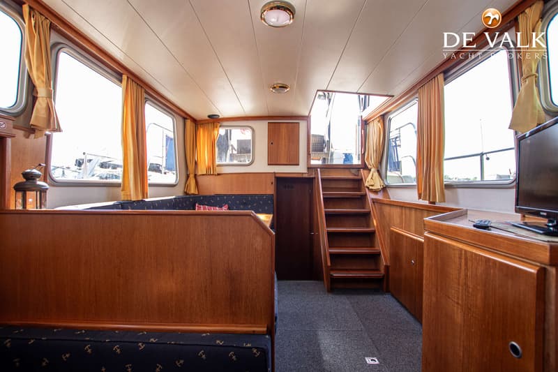 Een interieur van een boot met houten lambrisering, ramen met gele gordijnen, een trap en een bank met ankerpatroon.