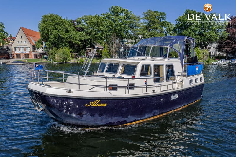 Een blauw-witte motorboot met de naam "Alexa" vaart op een waterweg met bomen en gebouwen op de achtergrond. — Pedro Marin 30