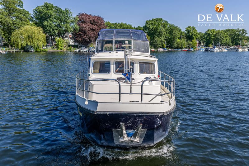 Een blauw-witte motorboot vaart op een meer met bomen langs de oever en een logo van "DE VALK YACHT BROKERS" rechtsboven.