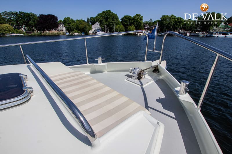 De foto toont het voordek van een boot met een ankerlier, reling en een gestreepte mat, met water en bebossing op de…