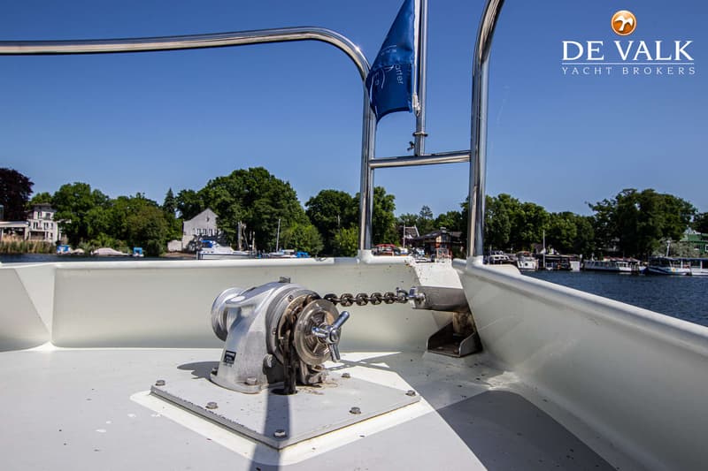 Een ankerlier met ketting op het voordek van een boot, met een blauwe vlag en een waterlandschap op de achtergrond.