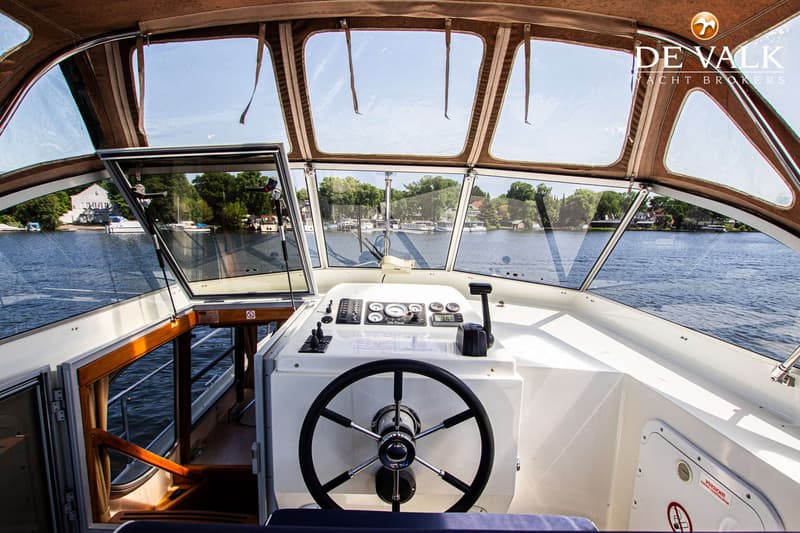 De foto toont de stuurstand van een boot met een stuurwiel, meters en bedieningselementen, uitkijkend over water.
