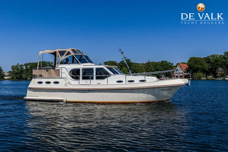 Een witte motorboot met een beige kap vaart op blauw water, met bomen en huizen op de achtergrond. — Gruno 36 Classic