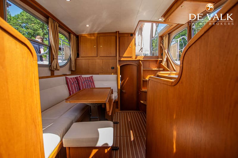 De foto toont het interieur van een boot met houten lambrisering, een bank met kussens, een tafel en een trap.
