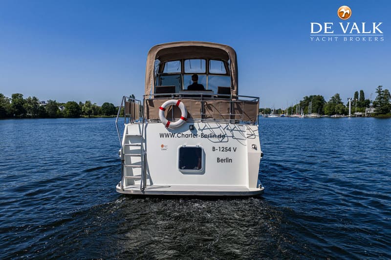 Een witte motorboot met een bruine kap vaart op een meer, met een zwemtrap en reddingsboei aan de achterzijde.