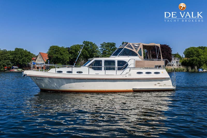 Een witte motorboot met de naam "Alexandra" vaart op een waterweg met beboste oevers en huizen op de achtergrond.