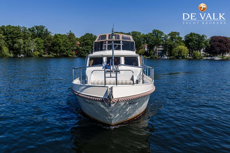 Een witte motorboot met de naam "Alexandra" op de boeg vaart op blauw water, omringd door groene bomen. — Gruno 36 Classic