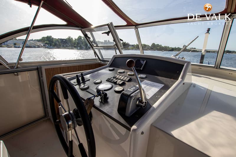 De foto toont de stuurstand van een boot met een stuurwiel, bedieningspaneel en een hendel, met uitzicht op water en…