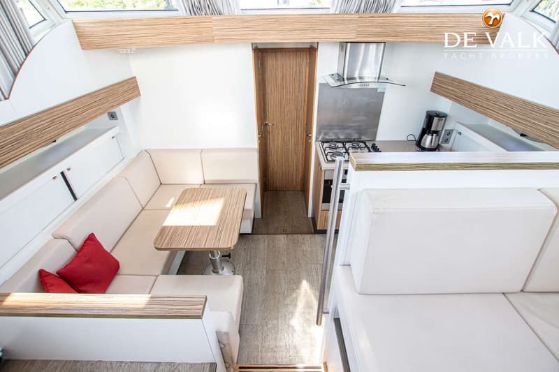 De foto toont een interieur van een boot met een zithoek, tafel, keukenblok met fornuis en koffiezetapparaat, en een houten…