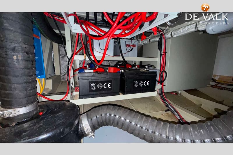 De foto toont een detail van de accu's en bedrading in een boot, met twee zwarte accu's op een plank en rode en zwarte…