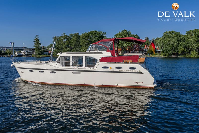 Een witte motorboot met rode bimini en Duitse vlag vaart op blauw water, met bomen en gebouwen op de achtergrond.