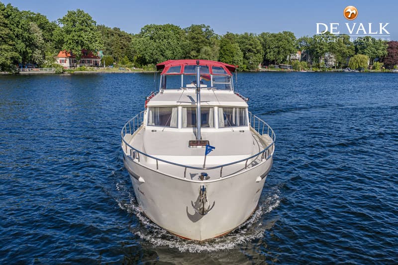 Een witte motorboot met een rood dak vaart op blauw water, met bomen en huizen op de achtergrond. — Linskens Catfish 46