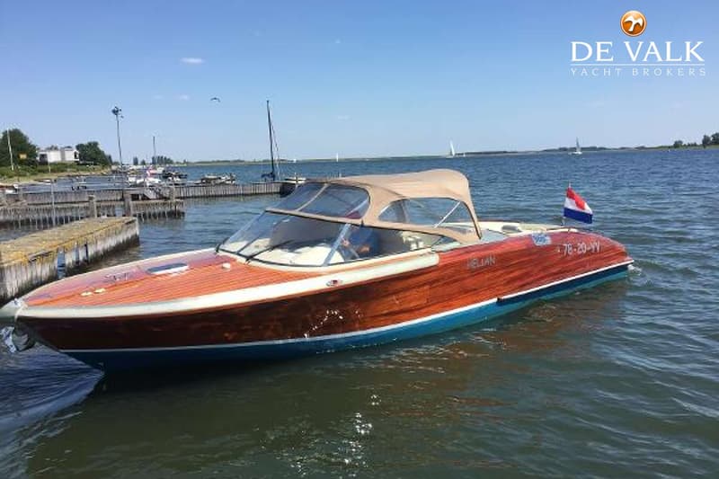 Een houten speedboot met een blauwe onderkant en een beige kap ligt in het water bij een steiger.