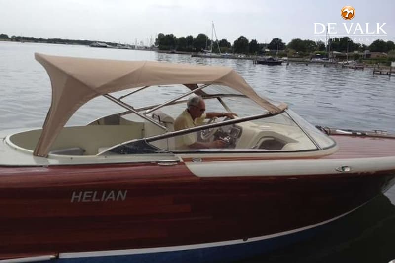 Een man met zonnebril zit aan het stuur van een bruin-witte boot met de naam "HELIAN" en een beige kap.