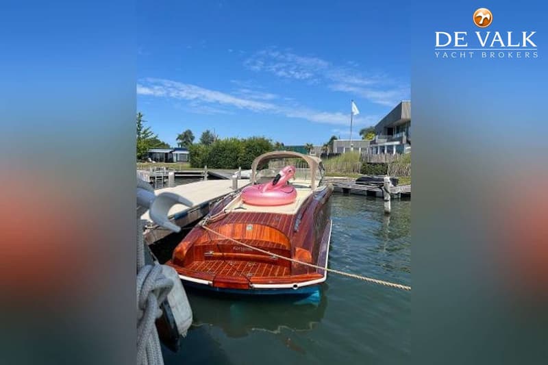 Een houten motorboot met een roze opblaasbare flamingo ligt aangemeerd aan een steiger onder een blauwe lucht.