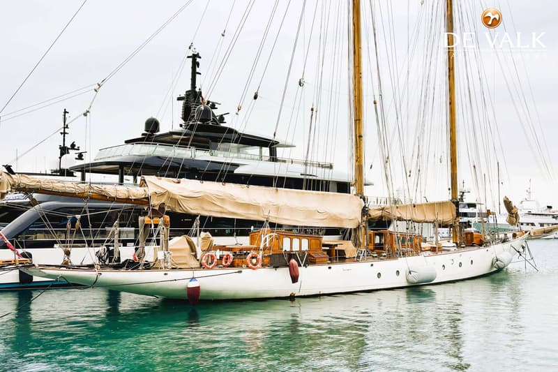 Een witte zeilboot met twee masten en opgerolde zeilen ligt in het water, met een donkere motorjacht op de achtergrond.