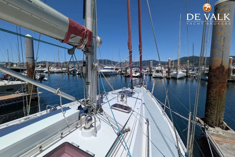 Een zeilboot ligt in een jachthaven met andere boten en bergen op de achtergrond onder een helderblauwe lucht.
