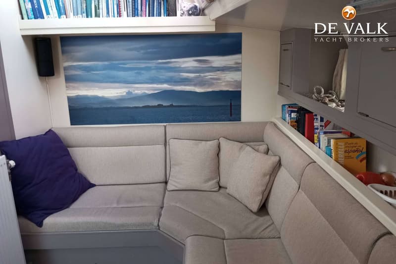 Een interieur met een L-vormige bank, kussens, boekenplanken en een ingelijste foto van een berglandschap.