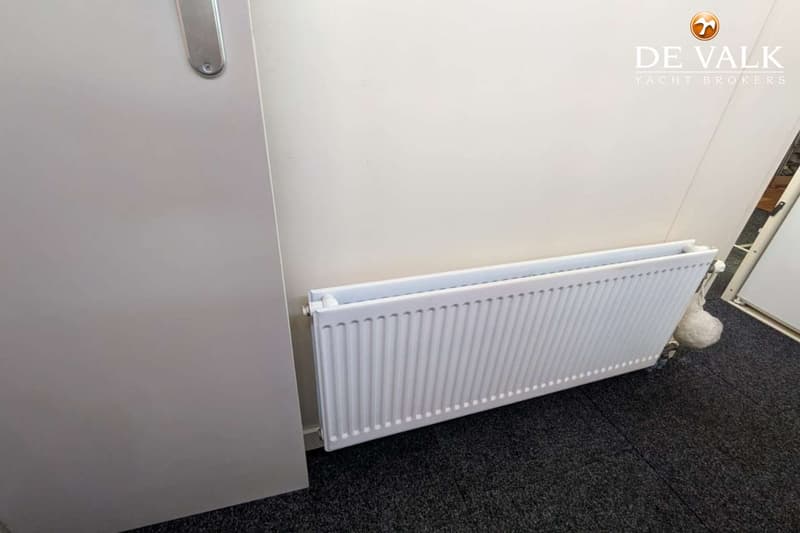 Een witte radiator is gemonteerd aan een lichte wand, met een donkergrijze vloerbedekking eronder.