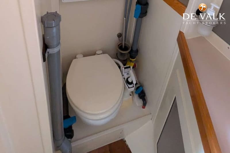 Een wit scheepstoilet met een beige bril en diverse leidingen eromheen, in een kleine ruimte.