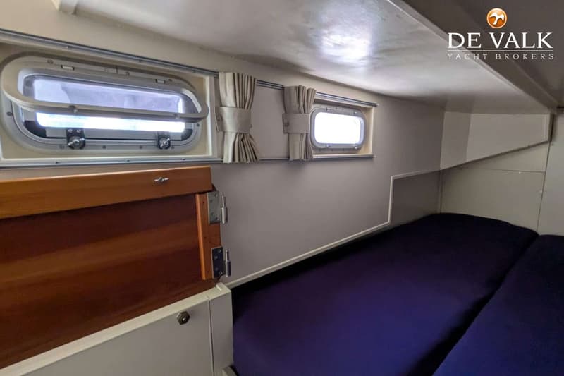 De foto toont een kajuit met twee patrijspoorten, gordijnen, een houten kast en twee bedden met donkerblauwe matrassen.