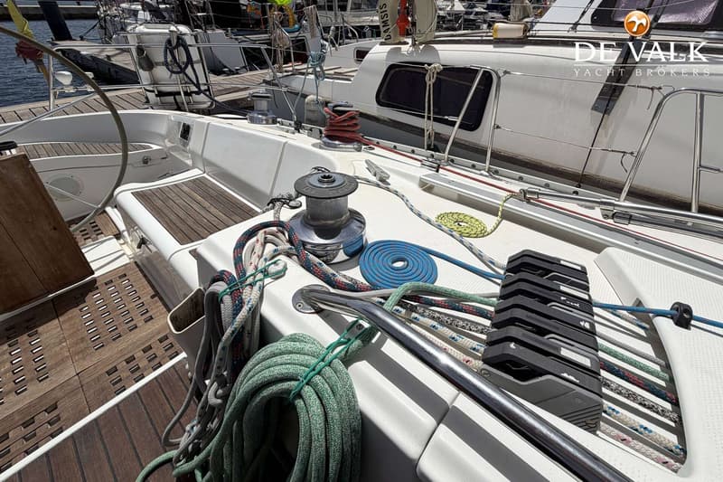 Een dek van een zeilboot met diverse touwen, lieren en een houten vloer, met een andere boot op de achtergrond.