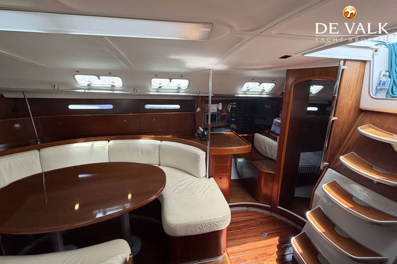De foto toont het interieur van een boot met een houten vloer, een U-vormige bank met crèmekleurige kussens en een ovale…